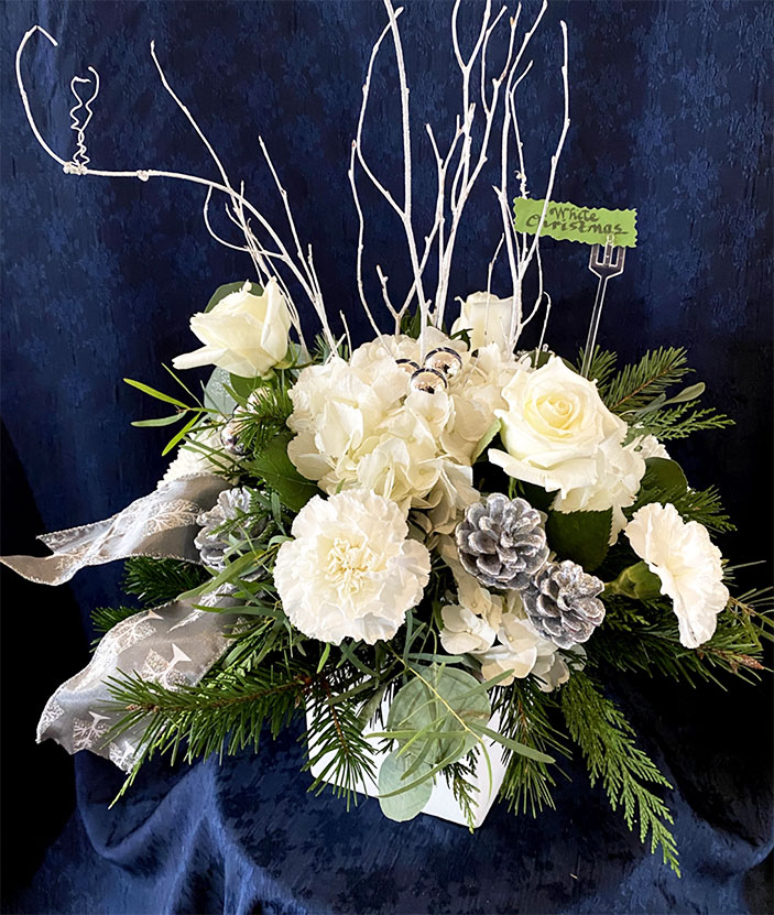 White Christmas $79 White ceramic cube, greens to include cedar, fir, and eucalyptus, white flowers of roses, hydrangea, carnations, disbuds, silver glass balls and pine cones, and tails of ribbon.